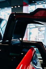 A red car with its gull-wing door open, set in a modern showroom with large windows allowing natural light to illuminate the space. The sleek, shiny surface of the car reflects the interior lighting and surroundings.