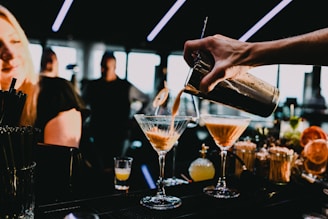 A bartender pours a cocktail into a martini glass with two other glasses visible on the countertop. A woman is in the foreground smiling, while the background features blurred figures and dim lighting. The bar area is stylishly arranged with various bottles and garnishes.