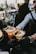 Two cocktails are elegantly presented on a bar counter, garnished with slices of passion fruit, fresh blackberries, and colorful fruit skewers. A bartender in a formal outfit and gloves is placing one of the glasses down, with a bustling bar atmosphere in the background.