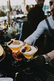 Two cocktails are elegantly presented on a bar counter, garnished with slices of passion fruit, fresh blackberries, and colorful fruit skewers. A bartender in a formal outfit and gloves is placing one of the glasses down, with a bustling bar atmosphere in the background.