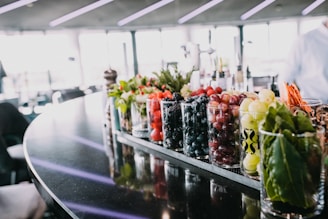 A countertop with a variety of fresh fruits, herbs, and vegetables in clear glass containers. The background is blurred, but it appears to be a modern indoor space with ample natural light coming through large windows. The fruits include strawberries, blueberries, blackberries, grapes, and apples. Herbs and leafy greens are also visible.