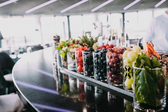 A countertop with a variety of fresh fruits, herbs, and vegetables in clear glass containers. The background is blurred, but it appears to be a modern indoor space with ample natural light coming through large windows. The fruits include strawberries, blueberries, blackberries, grapes, and apples. Herbs and leafy greens are also visible.