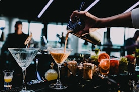 A bartender's hand, partially visible, is pouring a cocktail from a shaker into a glass on a bar counter. The bar counter holds several glasses, some with drinks garnished with fruit slices. The background shows a dimly lit environment with people and windows letting in natural light.