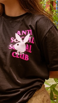 A person wearing a black t-shirt with pink lettering that reads 'ANTI SOCIAL SOCIAL CLUB' featuring a white rabbit design. The background includes greenery and foliage.
