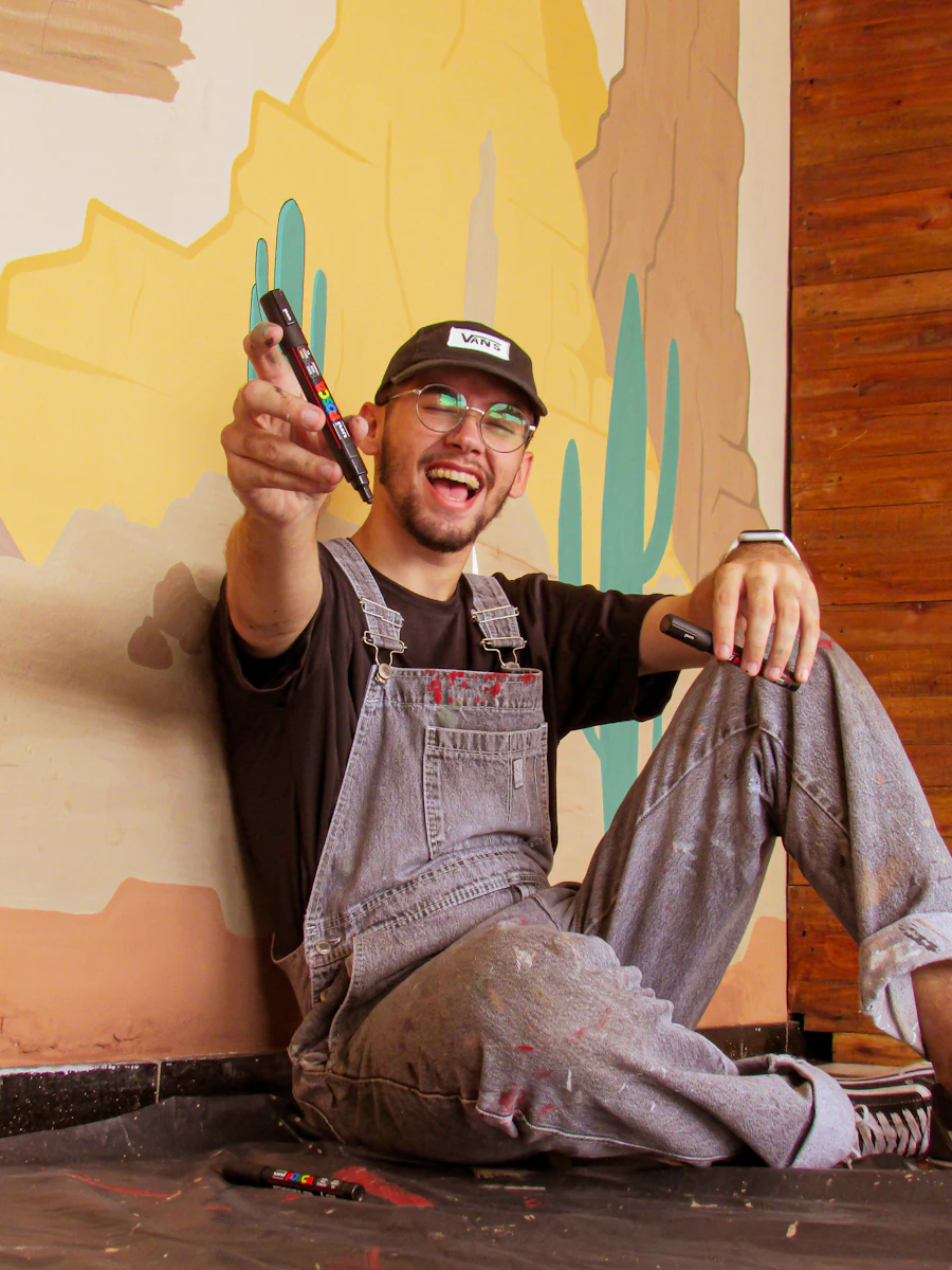 Mateo in painter's overalls with brushes in hand, in front of a saguaro mural