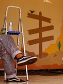 a man sitting on a step ladder painting a mural