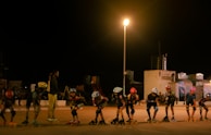 A group of young people are roller skating in a line under a bright streetlamp at night. They wear helmets and protective gear, and one adult appears to be coaching or instructing them. The background features some buildings and equipment, likely indicating a recreational area or park.
