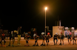 A group of young people are roller skating in a line under a bright streetlamp at night. They wear helmets and protective gear, and one adult appears to be coaching or instructing them. The background features some buildings and equipment, likely indicating a recreational area or park.