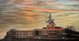 A large statue of a deity, surrounded by architectural structures with ornate details, is set against a sky painted with warm hues of orange and blue. The scene conveys a sense of grandeur and spirituality.