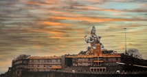 A large statue of a deity, surrounded by architectural structures with ornate details, is set against a sky painted with warm hues of orange and blue. The scene conveys a sense of grandeur and spirituality.
