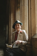 A person wearing a vintage-style outfit, including a green hat, a beige jacket, and a red floral shirt, is sitting by a stone window ledge. They are holding a book and smiling slightly, with a serene and intellectual atmosphere surrounding them. The background features stone archways and a dimly lit space, adding to the historic or academic feel.
