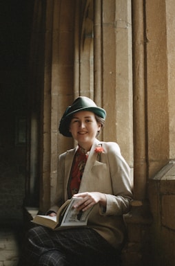 A person wearing a vintage-style outfit, including a green hat, a beige jacket, and a red floral shirt, is sitting by a stone window ledge. They are holding a book and smiling slightly, with a serene and intellectual atmosphere surrounding them. The background features stone archways and a dimly lit space, adding to the historic or academic feel.
