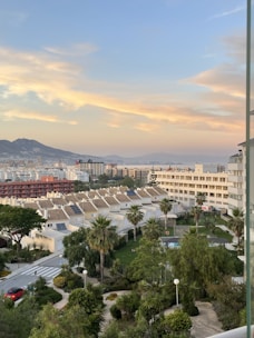 A cityscape featuring a row of white houses with sloped tile roofs, surrounded by lush greenery and palm trees. In the foreground, there's a garden area with trees and a small swimming pool. Further back, tall apartment buildings and a mountainous landscape can be seen under a sky with soft, orange-tinged clouds during sunset.