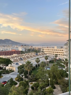 A cityscape featuring a row of white houses with sloped tile roofs, surrounded by lush greenery and palm trees. In the foreground, there's a garden area with trees and a small swimming pool. Further back, tall apartment buildings and a mountainous landscape can be seen under a sky with soft, orange-tinged clouds during sunset.
