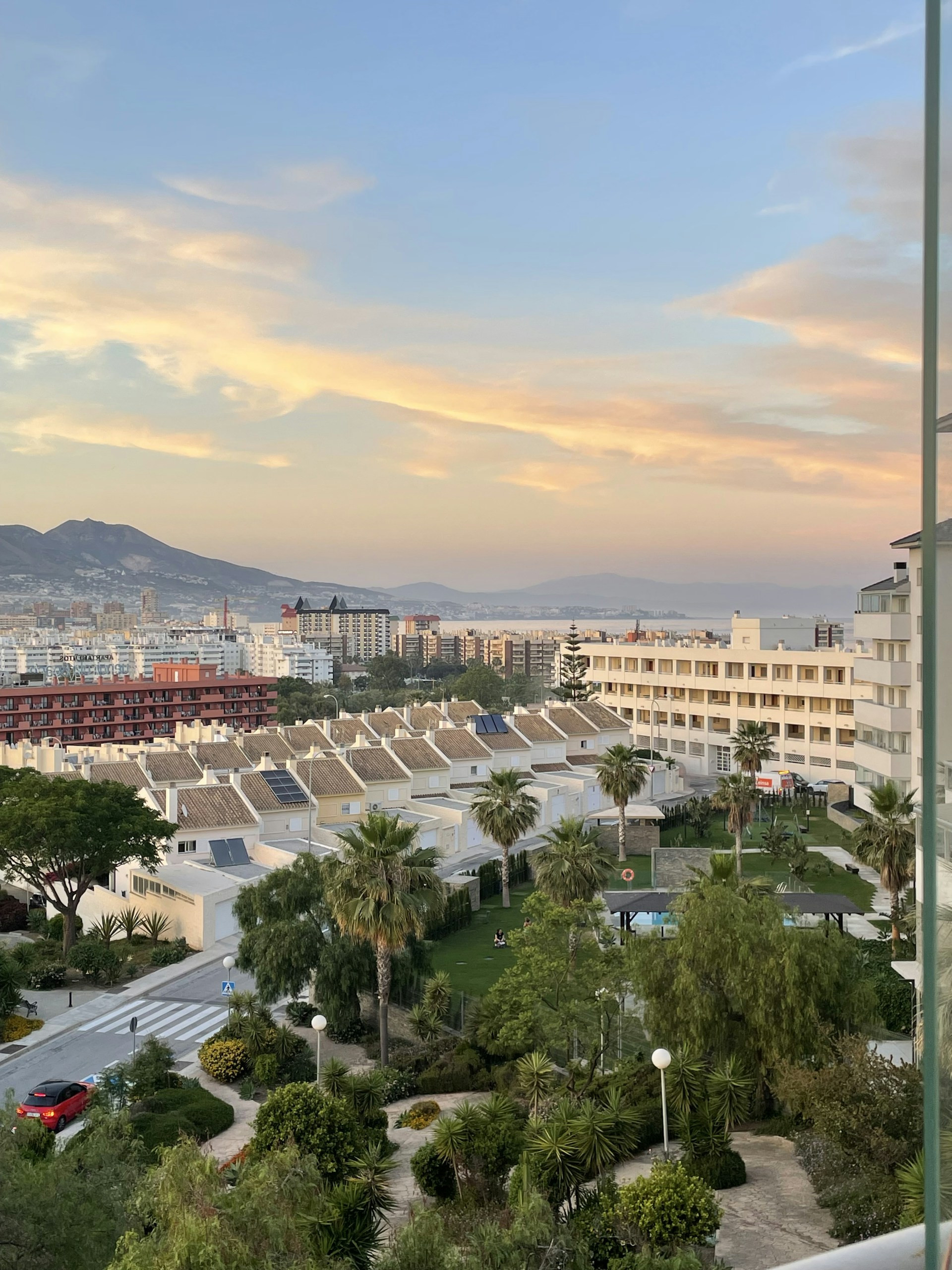 A cityscape featuring a row of white houses with sloped tile roofs, surrounded by lush greenery and palm trees. In the foreground, there's a garden area with trees and a small swimming pool. Further back, tall apartment buildings and a mountainous landscape can be seen under a sky with soft, orange-tinged clouds during sunset.