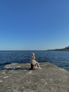 An elderly person sits on a stool at the edge of a concrete platform overlooking a calm body of water. The person appears to be wearing a light jacket and white shoes. The sky above is clear and blue, suggesting a peaceful, sunny day at a seaside location.