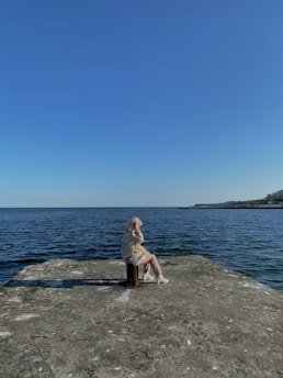An elderly person sits on a stool at the edge of a concrete platform overlooking a calm body of water. The person appears to be wearing a light jacket and white shoes. The sky above is clear and blue, suggesting a peaceful, sunny day at a seaside location.