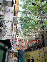 A narrow alley decorated with multiple international flags hanging between buildings. The alley has a vibrant atmosphere with green trees above and colorful flowers strung alongside the flags. There's a yellow wall mural on the right, depicting silhouettes of plants and creatures. A small cafe sign is visible, along with a money changer booth.