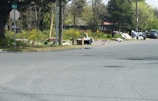 A street corner with scattered debris, discarded furniture, and litter. There is a stop sign on the left and a street sign mentioning '149 St S'. Trees and grass are present in the background, and there is a black SUV parked on the right side of the road.