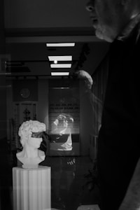 A black and white photo shows a reflective surface displaying the scene of a room. A bust of a classical statue is prominently displayed on a pedestal in the foreground. In the background, there's a man whose profile is partially visible, adding a sense of depth. The reflection captures a person holding a camera, creating a layered effect. The environment appears modern with ceiling lights and a clock on the wall.