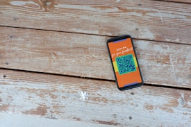 A smartphone with a colorful screen displaying the text 'scan me for your feedback' and a QR code lies on a wooden surface. The planks of wood have a rustic look with visible grain and some peeling paint.