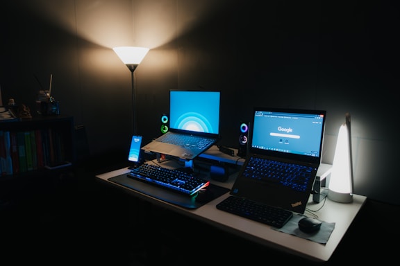 A dimly lit room features a desk with two open laptops side by side, each displaying colorful screens. A smartphone is positioned upright next to the laptops. The setup is accompanied by a keyboard and a mouse on a mouse pad. Two speakers are visible behind the laptops, adding to the immersive setup. The lighting is soft, provided by a floor lamp on the left and a table lamp on the right, enhancing the cozy atmosphere. Books are lined up on a shelf in the background.