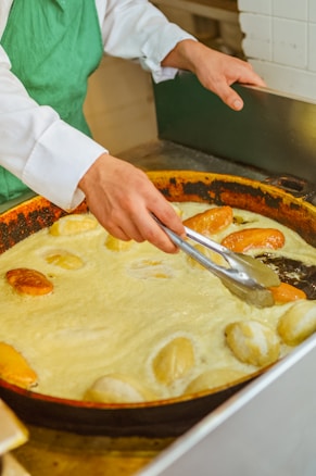 A person wearing a green apron is using tongs to fry bread rolls in a large pan filled with hot oil. The pan is situated on a stovetop, and the bread rolls are golden brown, indicating they are cooking.