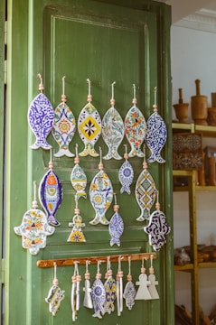 A wooden door painted green is adorned with an assortment of handcrafted ceramic ornaments. The ornaments, shaped like fish and other geometric forms, are intricately decorated with vibrant colors and floral patterns. In the background, shelves filled with wooden items like mortars add a rustic touch to the setting.