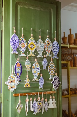 A wooden door painted green is adorned with an assortment of handcrafted ceramic ornaments. The ornaments, shaped like fish and other geometric forms, are intricately decorated with vibrant colors and floral patterns. In the background, shelves filled with wooden items like mortars add a rustic touch to the setting.