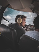 A person wearing headphones sits in the cockpit of a small aircraft, looking forward attentively. The airplane's dashboard is visible with various instruments and controls. Through the window, a partly cloudy sky can be seen.