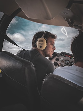 A person wearing headphones sits in the cockpit of a small aircraft, looking forward attentively. The airplane's dashboard is visible with various instruments and controls. Through the window, a partly cloudy sky can be seen.