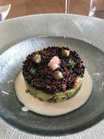A gourmet dish is artistically presented in a textured glass bowl, featuring layers of avocado and a topping of black and red caviar. Around the edges are small green herbs and capers. The dish is surrounded by a white creamy sauce.