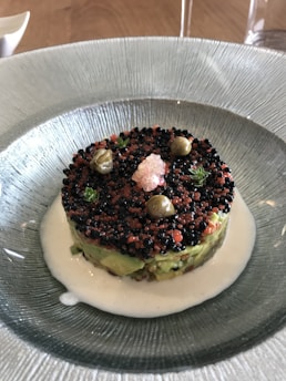A gourmet dish is artistically presented in a textured glass bowl, featuring layers of avocado and a topping of black and red caviar. Around the edges are small green herbs and capers. The dish is surrounded by a white creamy sauce.