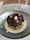 A gourmet dish is artistically presented in a textured glass bowl, featuring layers of avocado and a topping of black and red caviar. Around the edges are small green herbs and capers. The dish is surrounded by a white creamy sauce.
