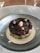 A gourmet dish is artistically presented in a textured glass bowl, featuring layers of avocado and a topping of black and red caviar. Around the edges are small green herbs and capers. The dish is surrounded by a white creamy sauce.