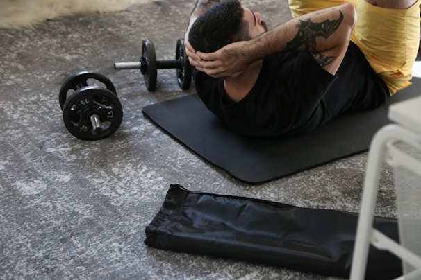 A man with tattoos performs abdominal exercises on a yoga mat indoors. A pair of adjustable dumbbells lies on the floor nearby, alongside a rolled-up exercise mat. The setting suggests a personal workout space, with an emphasis on strength training.