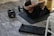 A man with tattoos performs abdominal exercises on a yoga mat indoors. A pair of adjustable dumbbells lies on the floor nearby, alongside a rolled-up exercise mat. The setting suggests a personal workout space, with an emphasis on strength training.