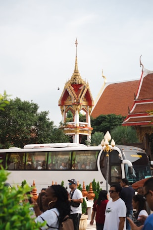 A traditional Thai temple structure with ornate, golden decorations and a pointed roof is visible. A large tour bus is parked nearby, filled with people, and there are many tourists walking around the area. The scene includes lush greenery and part of a temple roof.