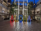Brightly illuminated letters spelling 'ALOHA' stand prominently in an open courtyard area surrounded by modern architectural elements. The letters are multicolored and adorned with small lights, creating a vibrant and welcoming atmosphere. Palm trees and string lights add to the tropical and festive ambiance.