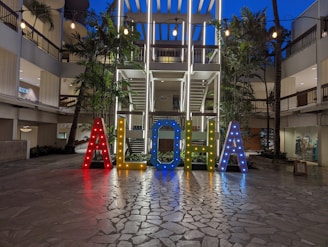 Brightly illuminated letters spelling 'ALOHA' stand prominently in an open courtyard area surrounded by modern architectural elements. The letters are multicolored and adorned with small lights, creating a vibrant and welcoming atmosphere. Palm trees and string lights add to the tropical and festive ambiance.