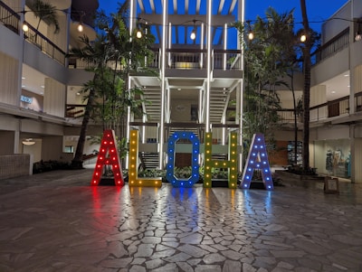 Brightly illuminated letters spelling 'ALOHA' stand prominently in an open courtyard area surrounded by modern architectural elements. The letters are multicolored and adorned with small lights, creating a vibrant and welcoming atmosphere. Palm trees and string lights add to the tropical and festive ambiance.
