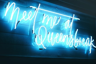 A neon sign with glowing blue and white text spelling out 'Meet me at Queensbreak' mounted on a wall with visible texture.