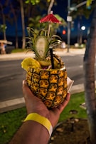A tropical drink served in a hollowed-out pineapple, garnished with a red cocktail umbrella, a slice of pineapple, and a straw. It is being held by a hand wearing a wristband, with a street scene and foliage in the background.