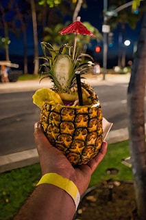 A tropical drink served in a hollowed-out pineapple, garnished with a red cocktail umbrella, a slice of pineapple, and a straw. It is being held by a hand wearing a wristband, with a street scene and foliage in the background.