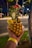 A tropical drink served in a hollowed-out pineapple, garnished with a red cocktail umbrella, a slice of pineapple, and a straw. It is being held by a hand wearing a wristband, with a street scene and foliage in the background.