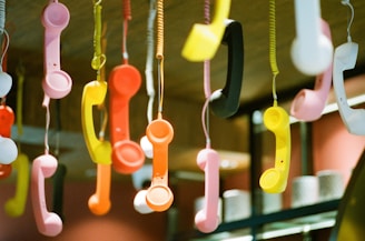 Hanging colorful retro telephone handsets are suspended from the ceiling by cords. The handsets are in various bright colors, including pink, yellow, orange, black, and white, creating a vibrant and eclectic display.
