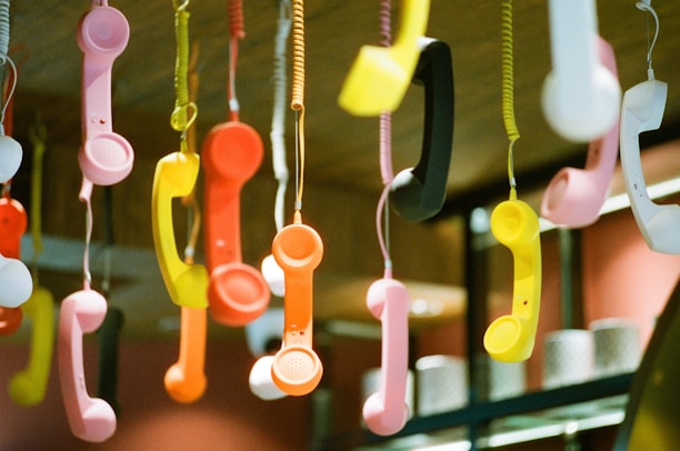 Hanging colorful retro telephone handsets are suspended from the ceiling by cords. The handsets are in various bright colors, including pink, yellow, orange, black, and white, creating a vibrant and eclectic display.