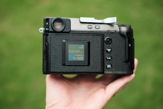 A digital camera held in a person's hand, showing the back view with a screen displaying settings information. The camera's buttons are visible, and it appears to be being used outdoors, indicated by the blurred green background.