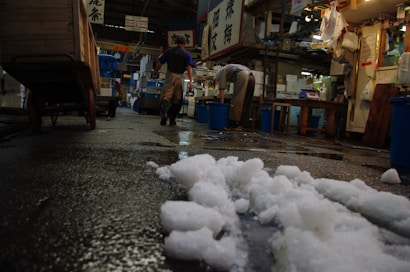 A bustling indoor market scene with wet floors and scattered piles of ice. Two workers wearing boots and aprons are busy at work, surrounded by various supplies and signage in a language with characters. The environment appears industrious and cluttered, containing ample storage boxes and equipment.