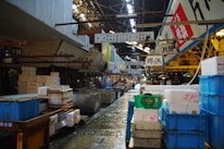 A bustling indoor market scene featuring a variety of crates and containers used for storage and transport. The market is filled with signs written in Japanese, hanging from above, adding to the authenticity and cultural vibe of the setting. The floor is wet, suggesting recent activity or cleaning. Workers are seen in the midground, busy with their tasks in this organized yet crowded space.